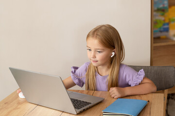 A smiling little girl with headphones conducts an online video call with a teacher using a laptop, communication of a small child with a tutor, online computer training, the concept of home learning