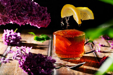 Black tea with lemon in a transparent glass cup, flying citrus slices and lilac branches and flowers