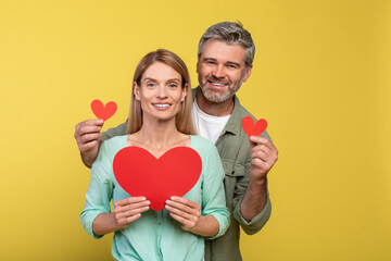 Love is in the air. Loving middle aged spouses with red paper hearts in hands posing over yellow studio background