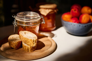 Still life. Slices of whole grain bread on a wooden board, a jar with homemade jam and ripe apricots on a white table