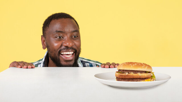 African Guy Looking At Burger On Table Over Yellow Background