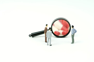 Three miniature figures of people look through a magnifying glass at medical capsules.