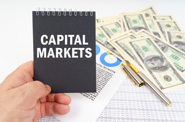 In the man's hand is a notepad with the inscription - CAPITAL MARKETS, in the background a pen and dollars