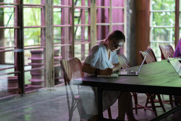 Young woman wearing glasses making notes, studying remotely in cafe, female student searching information in internet on laptop, watching webinar, learning foreign language online. Distance education
