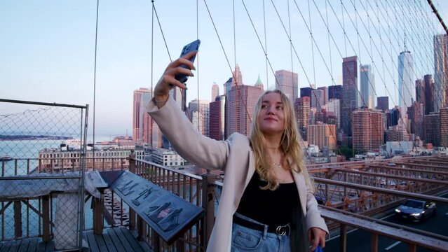 Beautiful young woman stands against the railing with business district in the background. Female tourist taking selfies at iconic Brooklyn Bridge. High quality 4k footage