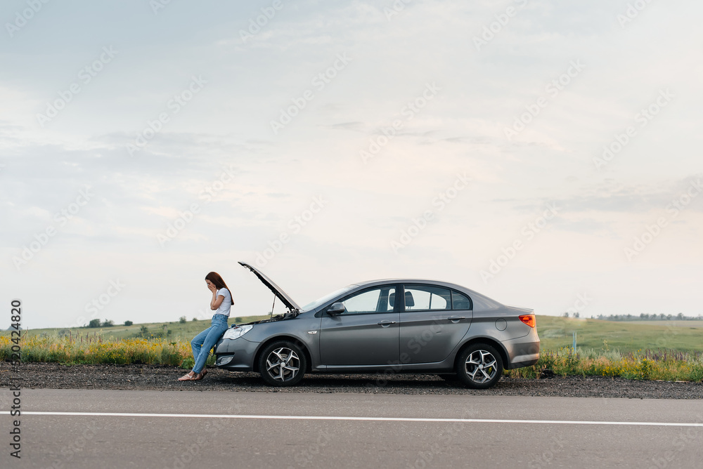 Wall mural A young girl stands near a broken car in the middle of the highway during sunset and tries to call for help on the phone and start the car. Waiting for help. Car service. Car breakdown on the road.