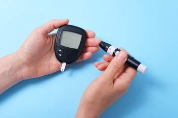 Woman using glucometer, checking blood sugar level. Diabetes concept on blue background. The hand holds the glucometer and pricks the finger with a lancet.