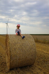 A modern village, hay bale on the field after the harvest on the background of wind turbine, windmill. The child is sitting on hay bale. Rural holiday.