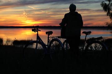 Fototapeta na wymiar Silhouette of a person with a bicycle standing on the high shore of the lake looking at the sunset back view at dusk