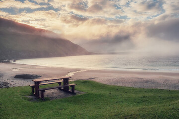 Table and benches for tourist on a grass with stunning view on Keem bay and beach early in the morning. Low clouds and fog over ocean and mountain. Ireland. Famous travel area. Irish landscape.