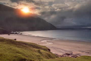 Keem bay and beach at sunrise. Dramatic dark cloudy sky low over ocean and mountain. Sun rising over a ridge. Calm and moody nature scene. County Mayo, Ireland. Irish landscape. Famous tourist area
