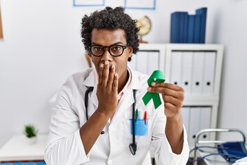African doctor man holding support green ribbon covering mouth with hand, shocked and afraid for...