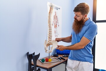 Young redhead man wearing physiotherapist uniform pointing to anatomical model of vertebral column...