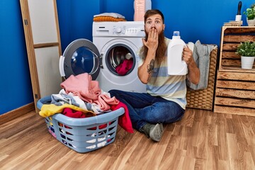 Redhead man with long beard doing laundry holding detergent bottle covering mouth with hand,...