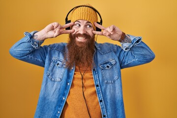 Caucasian man with long beard listening to music using headphones doing peace symbol with fingers...