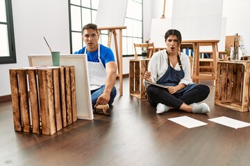 Young hispanic couple at art studio scared and amazed with open mouth for surprise, disbelief face
