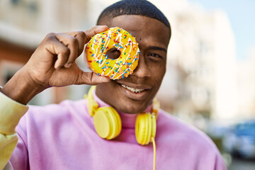 Young african american man smiling happy holding doughnut over eye at the city.