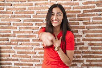 Young teenager girl standing over bricks wall laughing at you, pointing finger to the camera with hand over body, shame expression