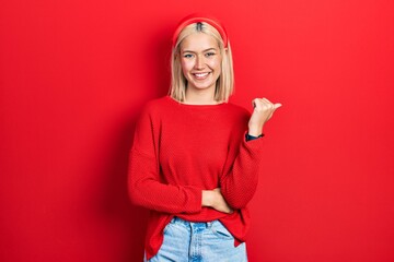 Beautiful blonde woman wearing casual red sweater smiling with happy face looking and pointing to...