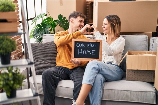 Young man and woman couple doing heart symbol holding blackboard sitting on sofa at new home