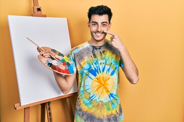 Young hispanic man standing drawing with palette by painter easel stand pointing with hand finger...