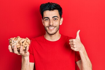 Young hispanic man holding peanuts smiling happy and positive, thumb up doing excellent and approval sign