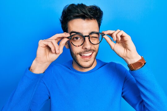 Young hispanic man wearing glasses winking looking at the camera with sexy expression, cheerful and happy face.