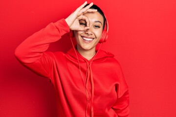 Fototapeta premium Beautiful hispanic woman with short hair listening to music using headphones doing ok gesture with hand smiling, eye looking through fingers with happy face.