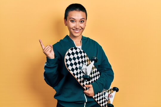 Beautiful hispanic woman with short hair holding skate smiling happy pointing with hand and finger to the side