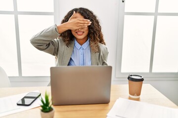 Beautiful hispanic business woman sitting on desk at office working with laptop smiling and laughing with hand on face covering eyes for surprise. blind concept.