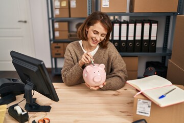 Young caucasian woman ecommerce business worker insert dollar on piggy bank at office