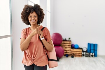 Naklejka na ściany i meble African american woman with afro hair holding yoga mat at pilates room with a big smile on face, pointing with hand finger to the side looking at the camera.