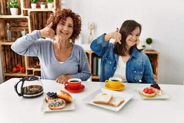 Family of mother and down syndrome daughter sitting at home eating breakfast smiling doing phone gesture with hand and fingers like talking on the telephone. communicating concepts.