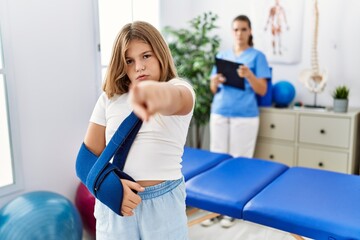 Blonde little girl wearing arm on sling at rehabilitation clinic pointing with finger to the camera and to you, confident gesture looking serious