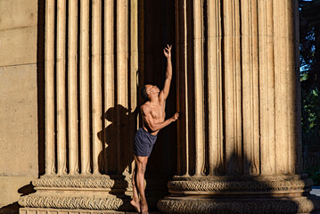 Beautiful ballet dancer standing against a column raising his hand as if letting go for a new...