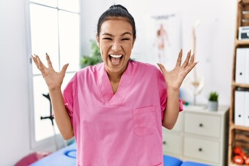 Young hispanic woman working at pain recovery clinic celebrating crazy and amazed for success with arms raised and open eyes screaming excited. winner concept