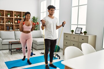 Young latin couple smiling happy training using elastic band and dumbbells at home.