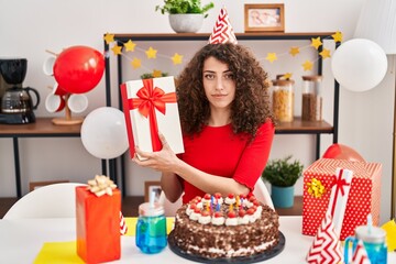 Hispanic woman with curly hair celebrating birthday with cake and present relaxed with serious expression on face. simple and natural looking at the camera.