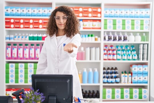 Hispanic woman with curly hair working at pharmacy drugstore smiling cheerful offering palm hand giving assistance and acceptance.
