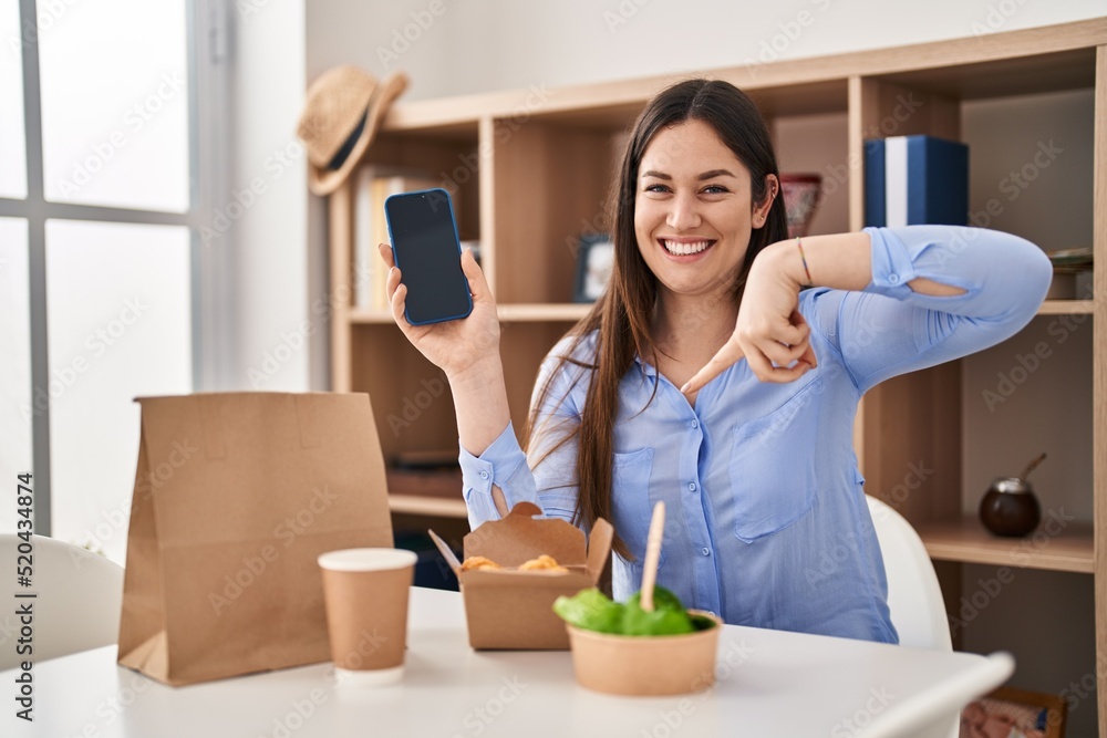 Wall mural young brunette woman eating take away food at home showing smartphone screen pointing finger to one 