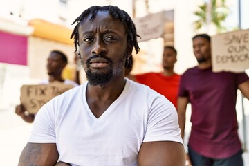 Young activist man with arms crossed gesture standing with a group of protesters holding banner protesting at the city.