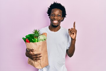 Young african american man holding paper bag with bread and groceries smiling with an idea or question pointing finger with happy face, number one