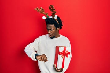 Young african american man wearing deer christmas hat holding gift looking at the watch time...