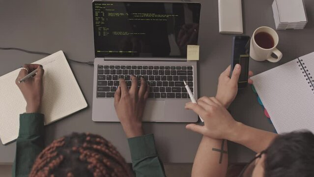 Top view slowmo of two diverse programmers working in team while developing new software writing program codes on laptop at office desk
