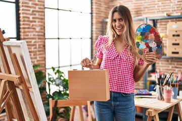 Young hispanic woman holding painter palette and art case clueless and confused expression. doubt concept.