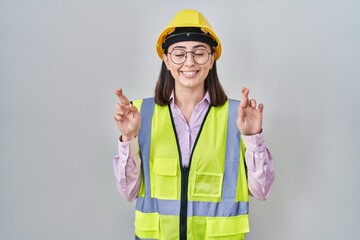 Hispanic girl wearing builder uniform and hardhat gesturing finger crossed smiling with hope and eyes closed. luck and superstitious concept.