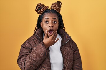 African woman with braided hair standing over yellow background looking fascinated with disbelief, surprise and amazed expression with hands on chin