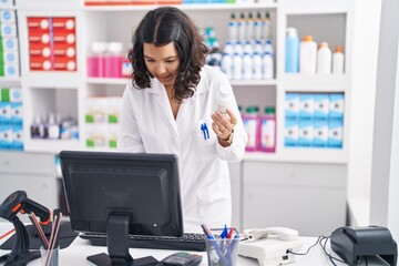 Young woman pharmacist holding pills bottle using computer at pharmacy