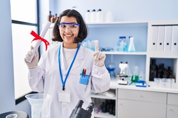 Young hispanic woman working at scientist laboratory holding degree smiling with an idea or question pointing finger with happy face, number one - obrazy, fototapety, plakaty