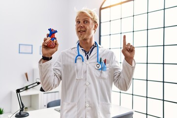 Young blond man wearing doctor uniform holding heart at clinic smiling with an idea or question...